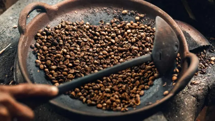 Grãos de café torrados em uma panela de metal escura sobre o fogo, com uma colher de metal.